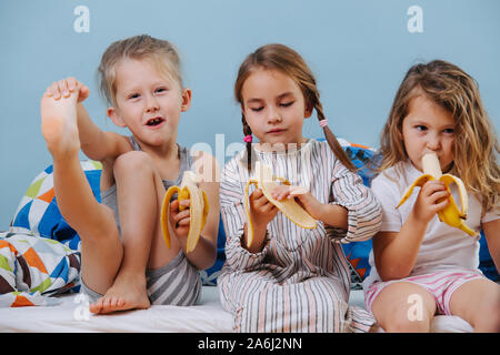 Drei süße kleine Kinder Nachtwäsche sitzen auf ungemachten Bett, essen Bananen Stockfoto