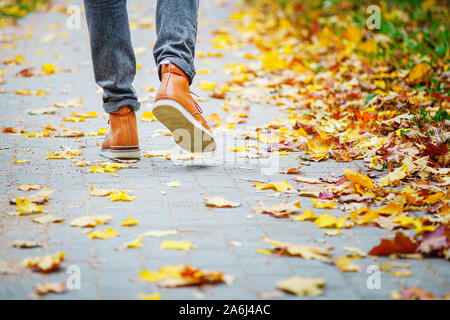 Zurück Blick auf die Füße von einem Mann im braunen Stiefel wandern entlang dem Bürgersteig mit Laub übersät. Der Begriff des Umsatzes der Jahreszeiten des Jahres Stockfoto