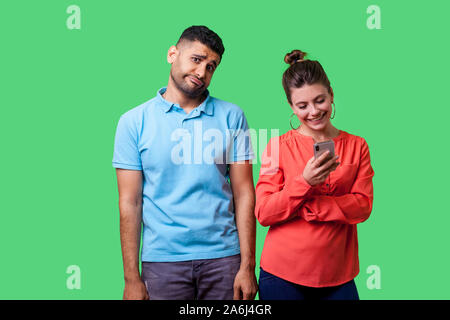 Portrait von lustigen umgekippt Mann in Freizeitkleidung mit Hände unten stehend, gelangweilt und genervt, wütend über Frau mit Smartphone, ihn zu ignorieren. isol Stockfoto