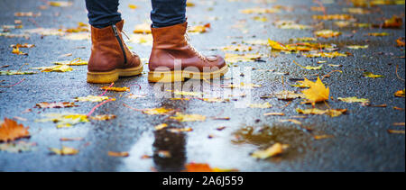 Spaziergang auf dem nassen Bürgersteig. Zurück Blick auf den Füßen einer Frau, die auf dem Asphalt mit Pfützen im Regen. Paar Schuhe auf glatter Straße in Th Stockfoto