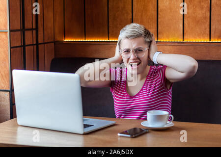 Möchten nicht hören! Portrait von attraktiven jungen Mädchen Freelancer in Rosa t-shirt sitzt im Cafe und Arbeiten am Laptop, die Ohren mit den Fingern Stockfoto