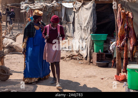 Opuwo, Namibia - 25. Juli 2019: Alte Frau in der traditionellen Kleidung der Herero und junge Schulmädchen in Schuluniform auf dem lokalen Markt in Opuwo zwischen Po Stockfoto