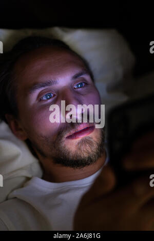 Junge stattlich und müde Mann mit einem Bart kann nicht schlafen und ist etwas aufpassen auf sein Handy in der Nacht. Stockfoto