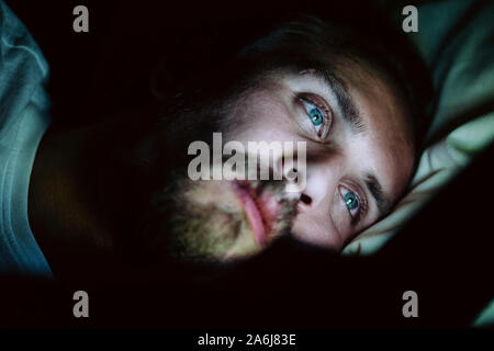 Bärtige Kaukasier Männlich ist etwas aufpassen auf seinem Mobiltelefon nachts im Bett. Er ist eindeutig sehr müde, aber kann nicht schlafen. Stockfoto
