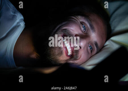 Bärtige glückliche junge Mann liegt in seinem Bett und lächelt, während etwas aufpassen auf seinem Telefon. Es ist eindeutig sehr spät in der Nacht. Stockfoto