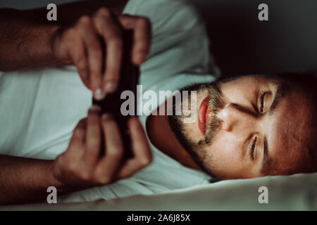 Junge glücklicher Mensch kann nicht schlafen und ist etwas aufpassen auf sein Handy in der Nacht. Weißes Licht aus das Telefon alle über sein Gesicht. Stockfoto