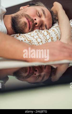 Telefon im Bett neben einem gutaussehenden jungen Mann, dass ist ein Nickerchen. Reflexion des Telefons zu sehen. Stockfoto