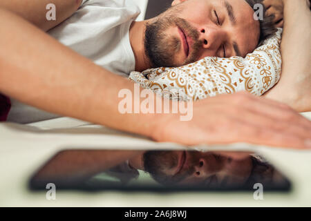 Telefon im Bett neben einem gutaussehenden jungen Mann, dass ist ein Nickerchen. Reflexion des Telefons zu sehen. Stockfoto