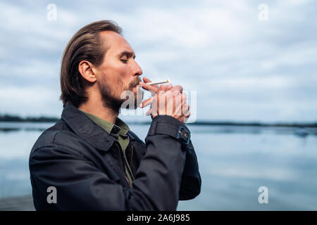 Foto eines Jungen bärtigen Mann Rauchen einer Zigarette. See und Wolken im Hintergrund. Stockfoto