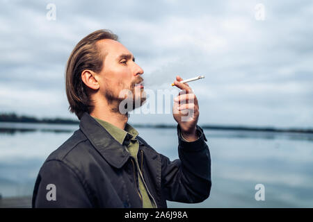 Foto eines Jungen bärtigen Mann Rauchen einer Zigarette. See und Wolken im Hintergrund. Stockfoto