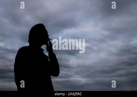 Dunkle Silhouette von einem gutaussehenden jungen Mann Rauchen einer Zigarette. Dunkle Wolken im Hintergrund sichtbar. Stockfoto