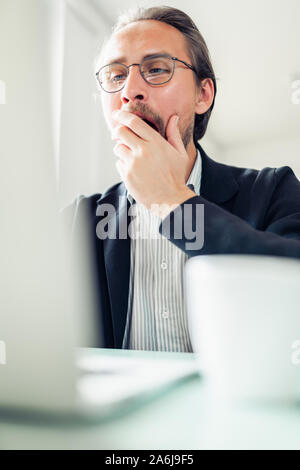 Müde jungen gutaussehenden Mann ist Gähnen während am Computer arbeiten. Kaffee Tasse sichtbar neben ihm. Stockfoto