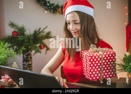 Eine schöne Frau in rotem Kleid hält eine Geschenkverpackung und Bestellung online Einkäufe auf Ihrem Laptop. Online Einkaufen für die Weihnachtsferien. Stockfoto
