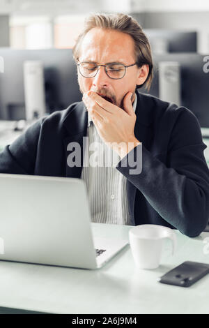 Müde jungen gutaussehenden Mann ist Gähnen während am Computer arbeiten. Tasse Kaffee und einem Mobiltelefon sichtbar neben ihm. Stockfoto