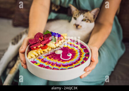 Junge Frau in ein mediterranes Frühstück auf Ihrem Sofa und mit ihrer Katze sitzt und isst Gesund tropisches Frühstück, smoothie Schüssel mit tropischen Stockfoto