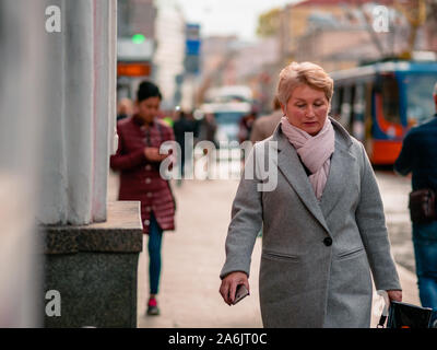 Moskau, Russland - Oktober 19, 2019: Ältere blonde Frau im Mantel hält in ihrer Hand Smartphone und Spaziergänge entlang der Straße. Das Telefon hat indispe werden Stockfoto