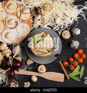 Vareniki gefüllt mit Pilzen und Kohl auf einem schwarzen Hintergrund mit Zutaten, Ansicht von oben, handgefertigt. Russisch, Ukrainisch, traditionelle Küche, culina Stockfoto