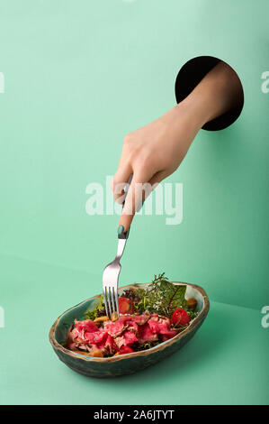 Weibliche Hand reicht für Roastbeef Salat durch das Loch in der grünen Wand. Essen Konzept in der neo-mint Farbe. Hand nimmt Nahrung durch das Loch. Rot Rindfleisch Salat Stockfoto