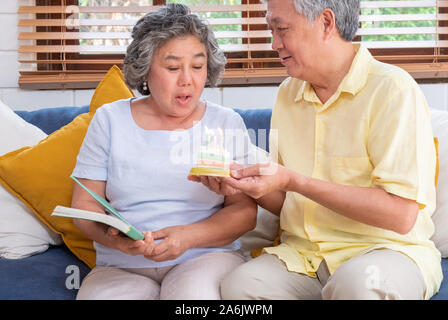 Asiatische älterer Mann überraschen ältere Frau mit Geburtstag Kuchen im Wohnzimmer zu Hause. Altern zu Hause Konzept. Stockfoto