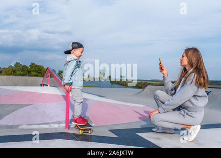 Frau Mama nimmt Bilder auf Telefon, Support Care training, kleinen Jungen 3-5 Jahre alten Sohn steht auf einem Skateboard, Züge im Sommer Stadt zu reiten, Herbst auf Stockfoto