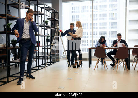Blond hübscher Kerl reden von Smart Phone, ein Gespräch mit Partnern, während seine Mitarbeiter im Hintergrund des Fotos brainstorming. nicht Stockfoto