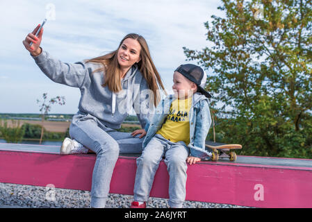 Frau Mama nimmt Bilder selfie Telefon kleinen Jungen 3-5 Jahre alten Sohn, sitzt das Ausruhen, Stadt für den Sommer Herbst Sportplatz glücklich lächelnde legere Kleidung Stockfoto