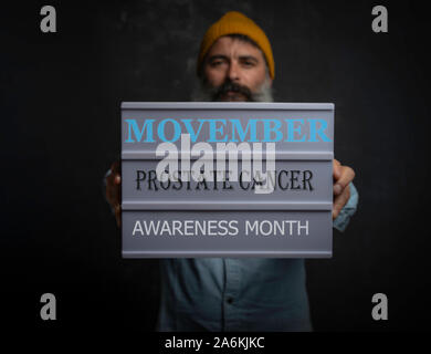 Movember Konzept. Portrait von unfocused bärtiger Mann mit einem Schild mit den Worten Movember, Prostatakrebs, ßtsein Monat im Vordergrund. Stockfoto