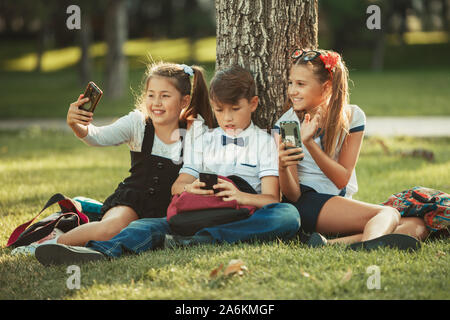 Drei Schule Freunde sitzen auf der Wiese unter einen Baum und spielen ihre Gadgets. Freunde nicht im Leben lieber das Telefon kommunizieren. Stockfoto