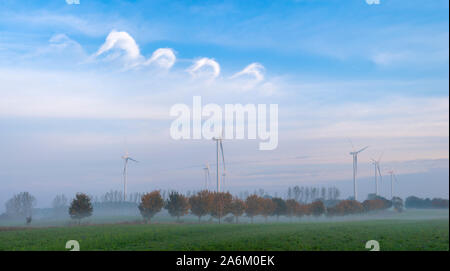 Schönen Herbst Landschaft. Die Reihe der Herbst Bäume im Nebel auf dem Feld, in der Ferne mit Windmühlen gegen den blauen Himmel Stockfoto