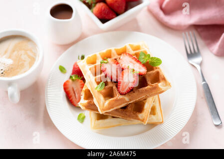 Belgische Waffeln mit Erdbeeren auf weißen Teller zum Frühstück serviert mit Schokoladensauce und Tasse Kaffee mit Sahne auf rosa Hintergrund Stockfoto