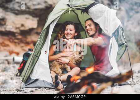 Paar trekker Sitzen im Zelt mit ihrem Haustier - glückliche Mann und Frau Spaß haben im Urlaub Camping rund um Rock Mountain neben dem Feuer Stockfoto