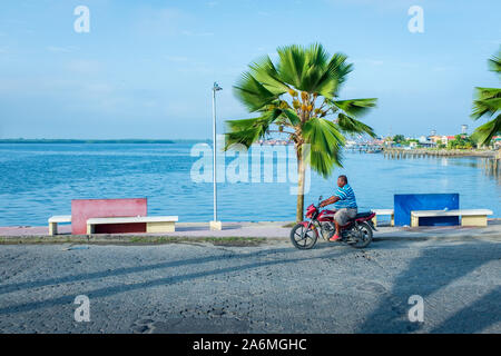 Tumaco, Nariño/Kolumbien, 17. Januar 2019: Schwarzer Mann Kreuzfahrt eine Brücke in der Nähe des Hafens in Motorrad, mit Palmen und Bänken Stockfoto