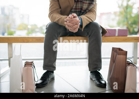 Geringer Teil der jungen stilvollen Mann in Jeans und Mantel sitzt auf der Bank im Trade Center Stockfoto