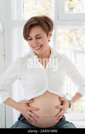 Stilvolle Mumie in weißem Hemd und blauer Jeans sitzt am Fenster. Schwangerschaft Konzept. Stockfoto