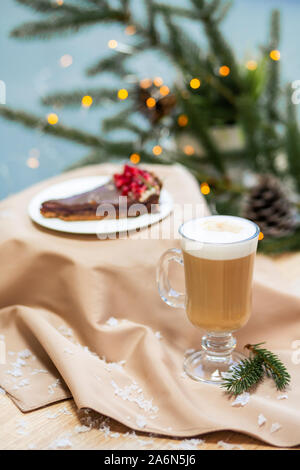 Köstliche frische festlichen morgen Cappuccino in einem Glas Schale und Kuchen Dessert auf dem hölzernen Tisch, Glühwürmchen und Zweige Fichte. Stockfoto