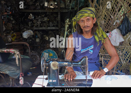 Ein Schneider mit seiner alten pedalgesteuerten Nähmaschine, der einen winzigen Platz vor einer Schrottwerkstatt einnimmt; Bhendi Bazar, Mumbai, Indien Stockfoto