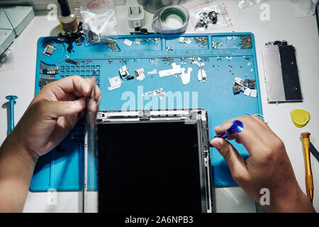 Handwerker mit Pinzette und Schraubendreher beim Entfernen der Bildschirm des Tablet-PC an seinem Schreibtisch, Ansicht von oben Stockfoto