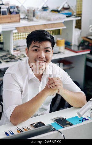 Fröhliche hübscher junger asiatischer Mann an Schreibtisch mit demontierten Smartphone sitzen Stockfoto