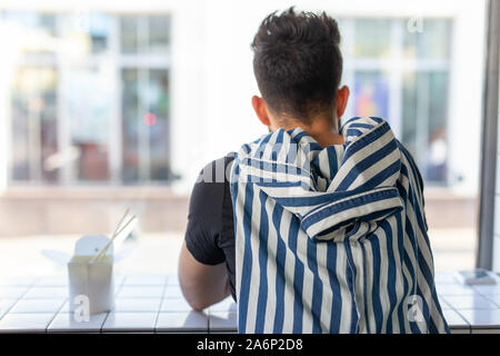 Hübscher junger hipster Kerl essen Chinesische Nudeln mit hölzernen Eßstäbchen in einem Cafe saßen und in das Fenster. Asiatische Küche Konzept Stockfoto