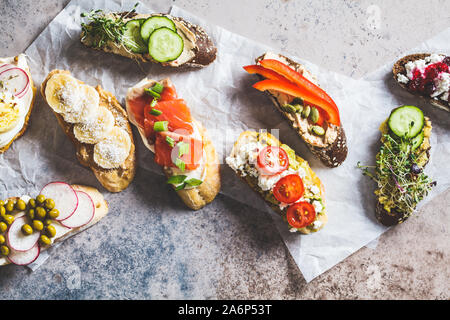 Toast mit verschiedenen Belägen auf grau-braunen Hintergrund geöffnet. Flach von Crostini mit Banane und Erdnussbutter, Pastete, Avocado, Lachs, Ei, Käse Stockfoto