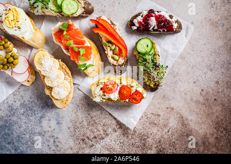 Toast mit verschiedenen Belägen auf grau-braunen Hintergrund geöffnet. Flach von Crostini mit Banane und Erdnussbutter, Pastete, Avocado, Lachs, Ei, Käse Stockfoto