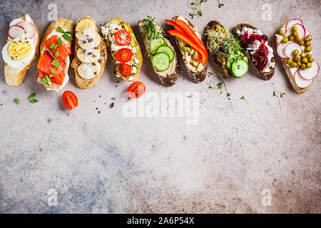 Toast mit verschiedenen Belägen auf grau-braunen Hintergrund geöffnet. Flach von Crostini mit Banane und Erdnussbutter, Pastete, Avocado, Lachs, Ei, Käse Stockfoto