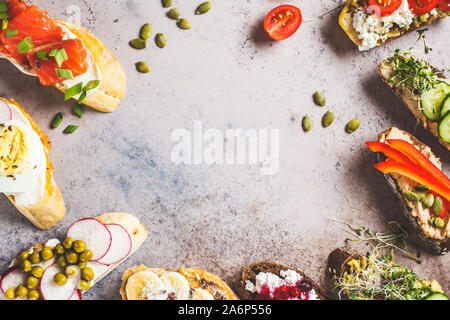 Toast mit verschiedenen Belägen auf grau-braunen Hintergrund geöffnet. Rahmen des Crostini mit Banane und Erdnussbutter, Pastete, Avocado, Lachs, Ei, Käse und Stockfoto