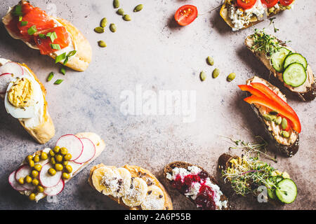 Toast mit verschiedenen Belägen auf grau-braunen Hintergrund, Kopieren offenen Raum. Rahmen des Crostini mit Banane und Erdnussbutter, Pastete, Avocado, Lachs, Ei Stockfoto