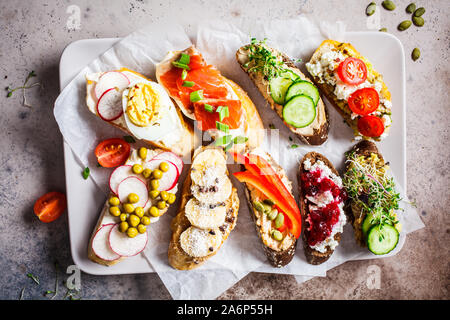 Toast mit verschiedenen Belägen auf grau-braunen Hintergrund geöffnet. Crostini mit Banane und Erdnussbutter, Pastete, Avocado, Lachs, Ei, Käse und Beeren. Stockfoto