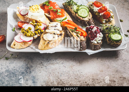 Toast mit verschiedenen Belägen auf grau-braunen Hintergrund, Kopieren offenen Raum. Crostini mit Banane und Erdnussbutter, Pastete, Avocado, Lachs, Ei, Käse ein Stockfoto