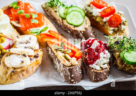 Toast mit verschiedenen Belägen auf grau-braunen Hintergrund geöffnet. Crostini mit Banane und Erdnussbutter, Pastete, Avocado, Lachs, Ei, Käse und Beeren. Stockfoto