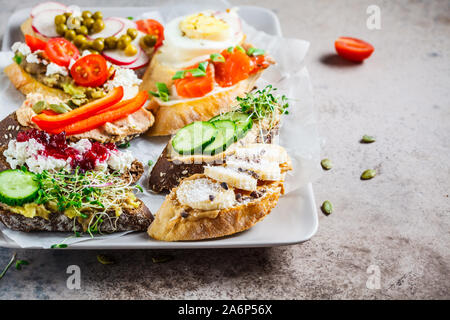 Toast mit verschiedenen Belägen auf grau-braunen Hintergrund, Kopieren offenen Raum. Crostini mit Banane und Erdnussbutter, Pastete, Avocado, Lachs, Ei, Käse Stockfoto