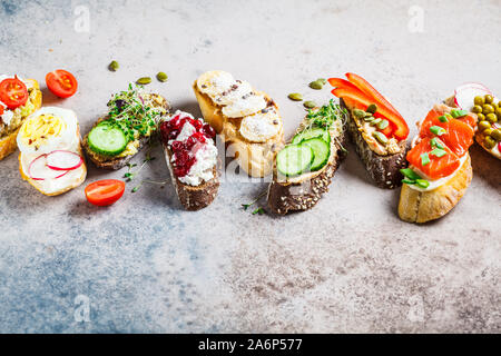 Toast mit verschiedenen Belägen auf grau-braunen Hintergrund, Kopieren offenen Raum. Flach von Crostini mit Banane und Erdnussbutter, Pastete, Avocado, Lachs, Stockfoto