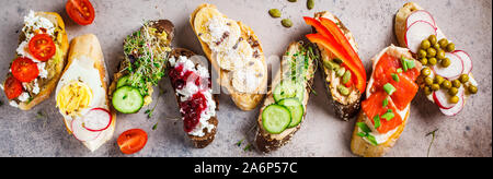 Toast mit verschiedenen Belägen auf grau-braunen Hintergrund geöffnet. Flach von Crostini mit Banane und Erdnussbutter, Pastete, Avocado, Lachs, Ei, Käse Stockfoto